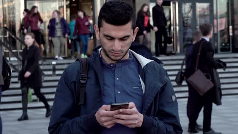 young middle eastern man using smartphone in city, people are walking behind