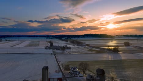 aerial hyperlapse of rolling clouds in dramatic sunset above rural landscape, sun beams reflect in frozen fields, traditional farm in america, winter nature time lapse