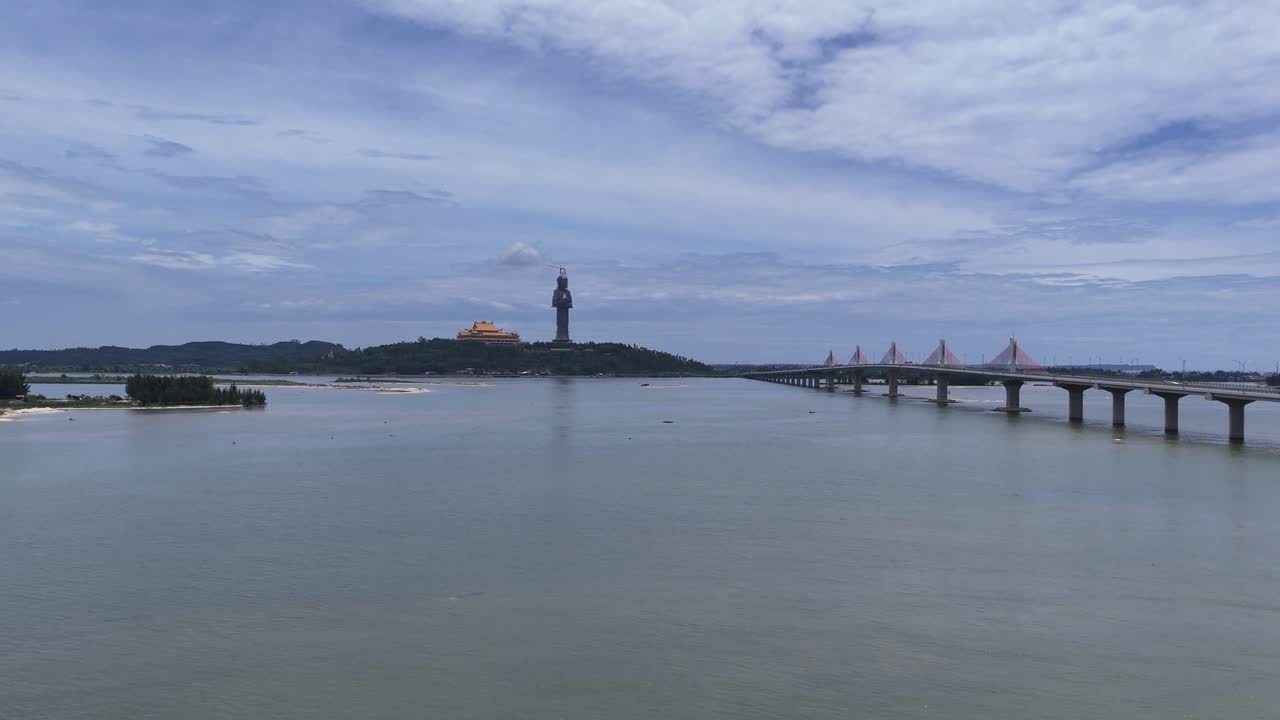 Aerial drone view of scenic river with long bridge stretching toward tall hilltop statue and golden temple, surrounded by water, sandbars, and lush countryside under bright cloudy sky