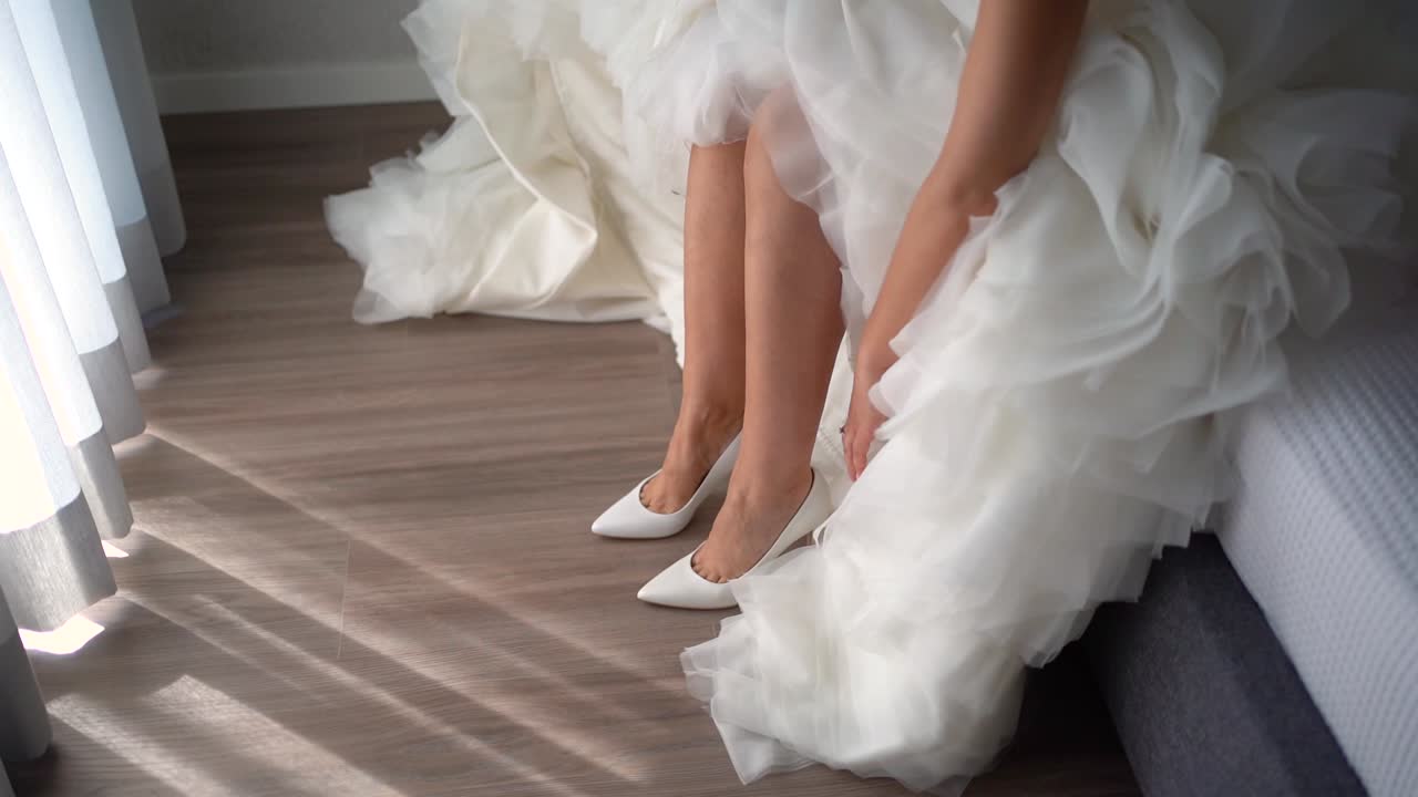 Bride in a white long elegant wedding dress puts on high-heeled white shoes