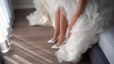 bride in a white long elegant wedding dress puts on high-heeled white shoes