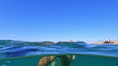 Half-underwater-rear-view-of-adult-man-fishing-and-swimming-in-blue-Adriatic-sea-with-diving-fins-along-trabocchi-traditional-platforms-in-Abruzzo,-Italy