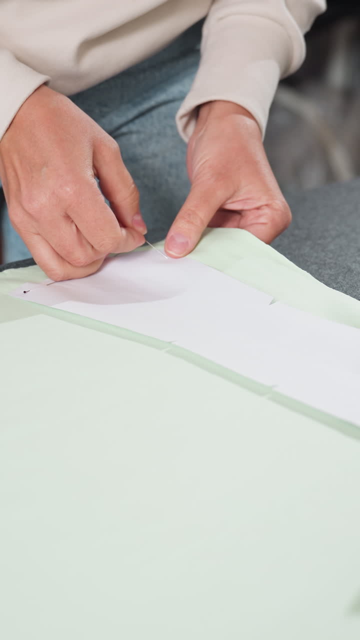 창백한 녹색 천을 따라 시접을 고정하는 손, 길게 정렬되어 핀으로 고정된 패턴 스트립, 옆에 놓인 나무 무게추, 꼼꼼한 기술과 디테일한 워크숍 집중력을 보여주는 클로즈업