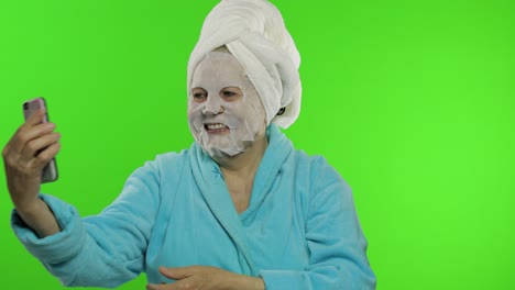 Grandmother-in-bathrobe,-face-mask.-Old-woman-making-selfies-using-mobile-phone