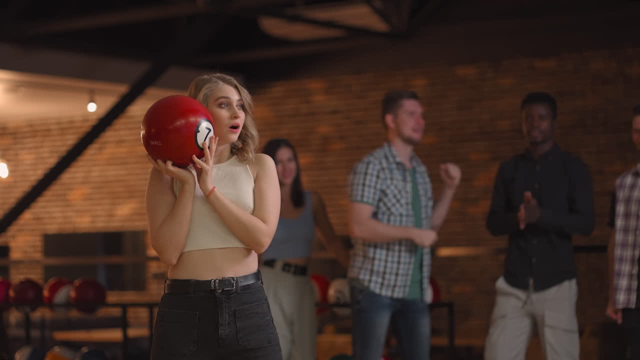 Portrait A young blonde woman makes the final throw with a bowling ball and wins the game with the support and joy of her friends of different nationalities. Rejoice and celebrate the victory