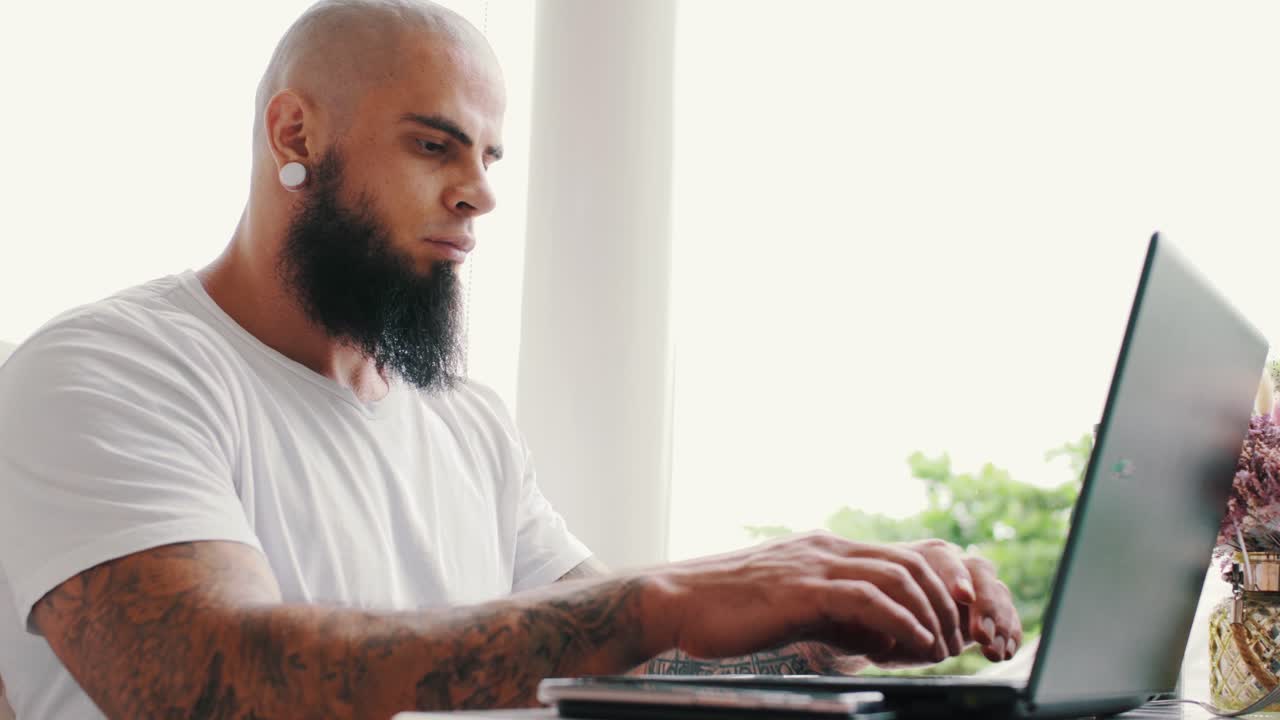 Handsome brutal young man with beard and tattoos is working on laptop near window in bright space