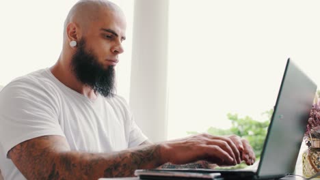 handsome brutal young man with beard and tattoos is working on laptop near window in bright space