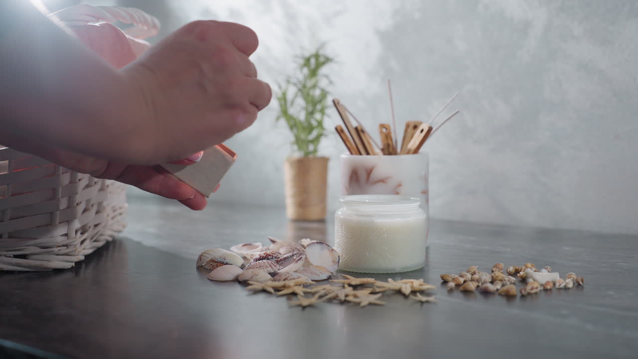Hand view of lady striking match to light white candle on marble table near arranged seashells and decorative items as bright light shines through window