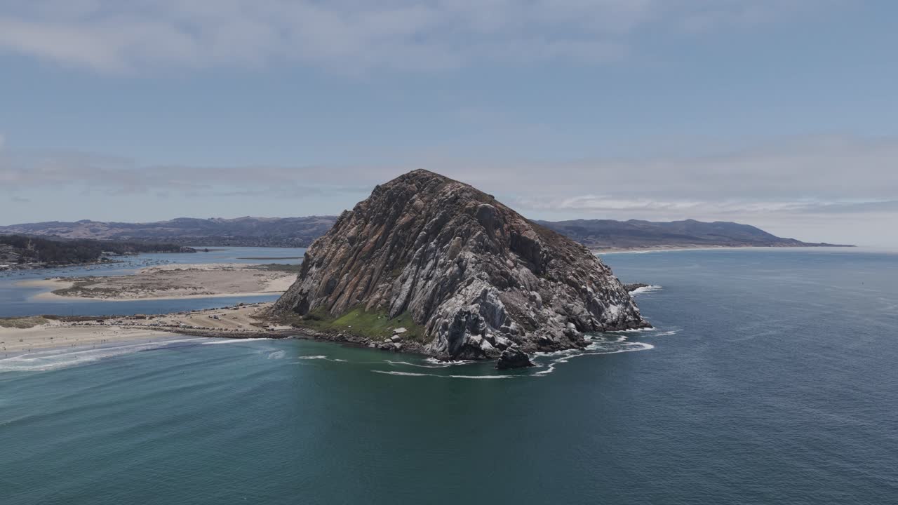 Wide drone shot flying over the Pacific Ocean toward Morro Rock in Morro Bay, California. The massive volcanic plug stands tall along the coast, surrounded by calm water and scenic shoreline