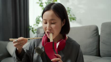 young asian woman in headphones eats noodles with wood sticks at home