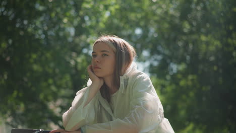 young woman in translucent raincoat leans thoughtfully on bicycle handlebars, resting her hand on her chin as sunlight gently filters through surrounding greenery