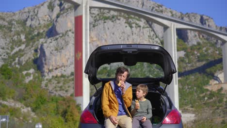 un hombre y su hijo están sentados y comiendo en su coche con el puente de moracica en el fondo. están tomando un viaje en coche a través de montenegro