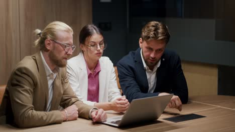 a brunette man in a blue jacket explains to his colleagues in the office how to perform this task correctly. meeting of colleagues in the office, a blond man in a light brown jacket and a brunette girl in a white suit sit in front of a laptop screen and communicate