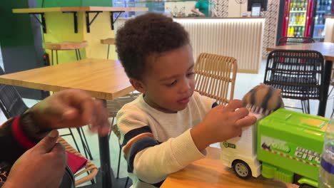 3-year-old-black-kid-playing-with-his-dinosaur-puppet-and-a-toy-truck-in-a-cafeteria-next-to-his-mother