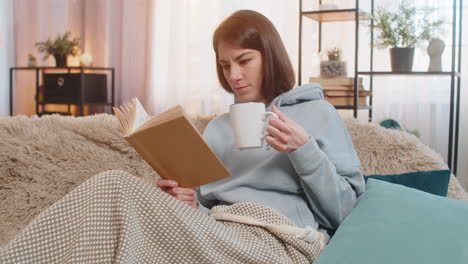 smiling young woman reading interesting book while having cup of hot chocolate on couch at home