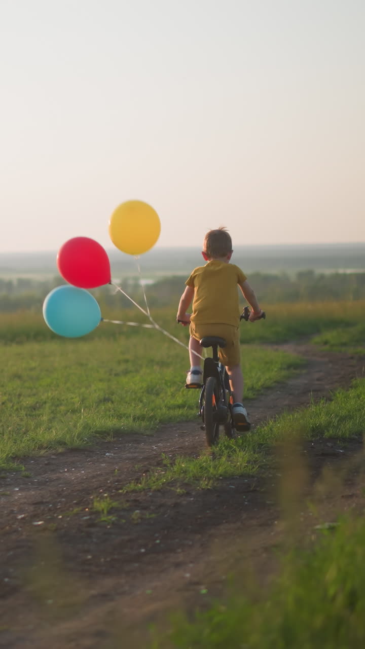 A young boy, dressed in yellow, rides his bicycle along a dirt path through a grassy field at sunset. Red, blue and yellow balloons are tied to the back of his bike. A car is visible in the background