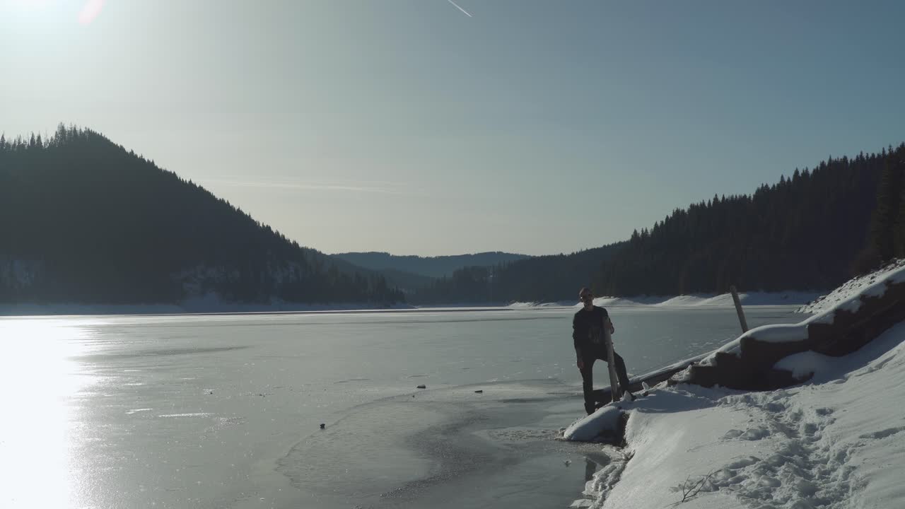 un hombre se para en la orilla del lago congelado en transilvania, rumania, sosteniendo un trozo de madera en un brillante día de invierno - plano general