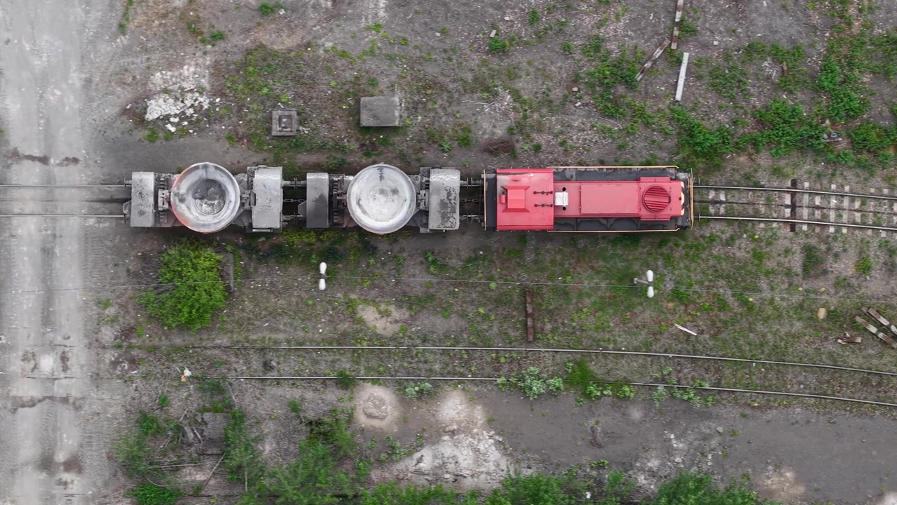 Una vista aerea cattura un treno industriale rosso con grandi vagoni merci su binari ferroviari paralleli, suggerendo trasporto pesante e industria.