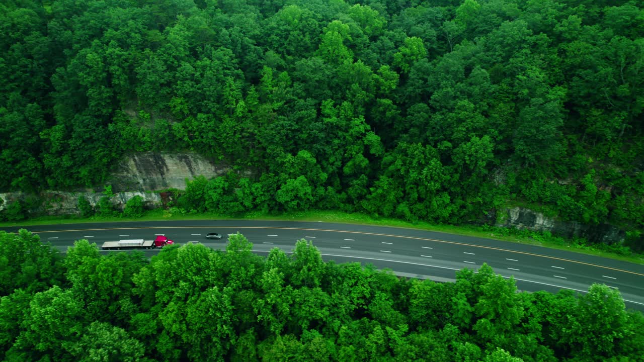 Red semi truck with flatbed trailer drives along forest highway past rocky cliff. Monteagle
Tennessee, USA.