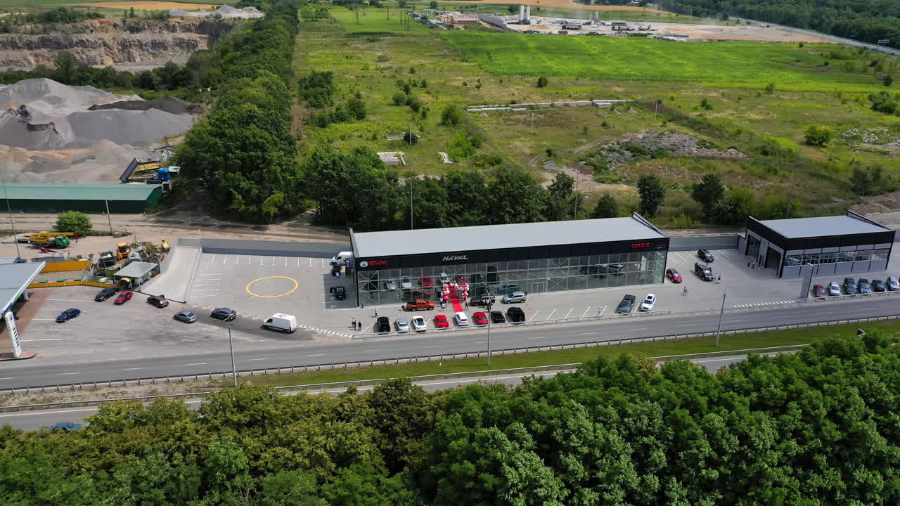 Flying closer to a newly-opened car dealership with automobiles at the entrance. Green field and industrial zone at backdrop. Top view.