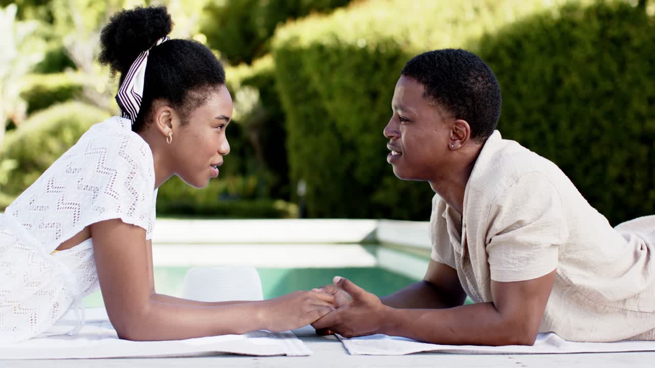 African American couple holding hands on towel at pool, man speaking, woman in white dress leaning