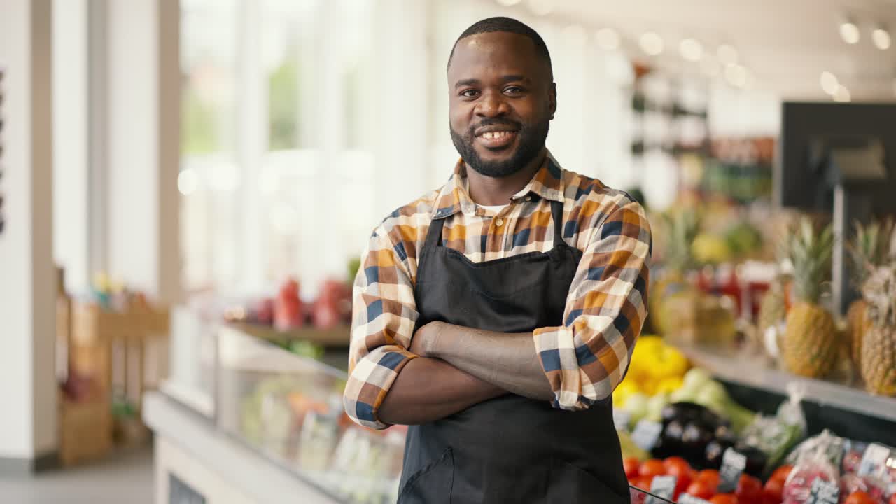 Portrait of a Black-skinned man in a plaid shirt and black apron folded his arms over his chest and looks at the camera in a supermarket