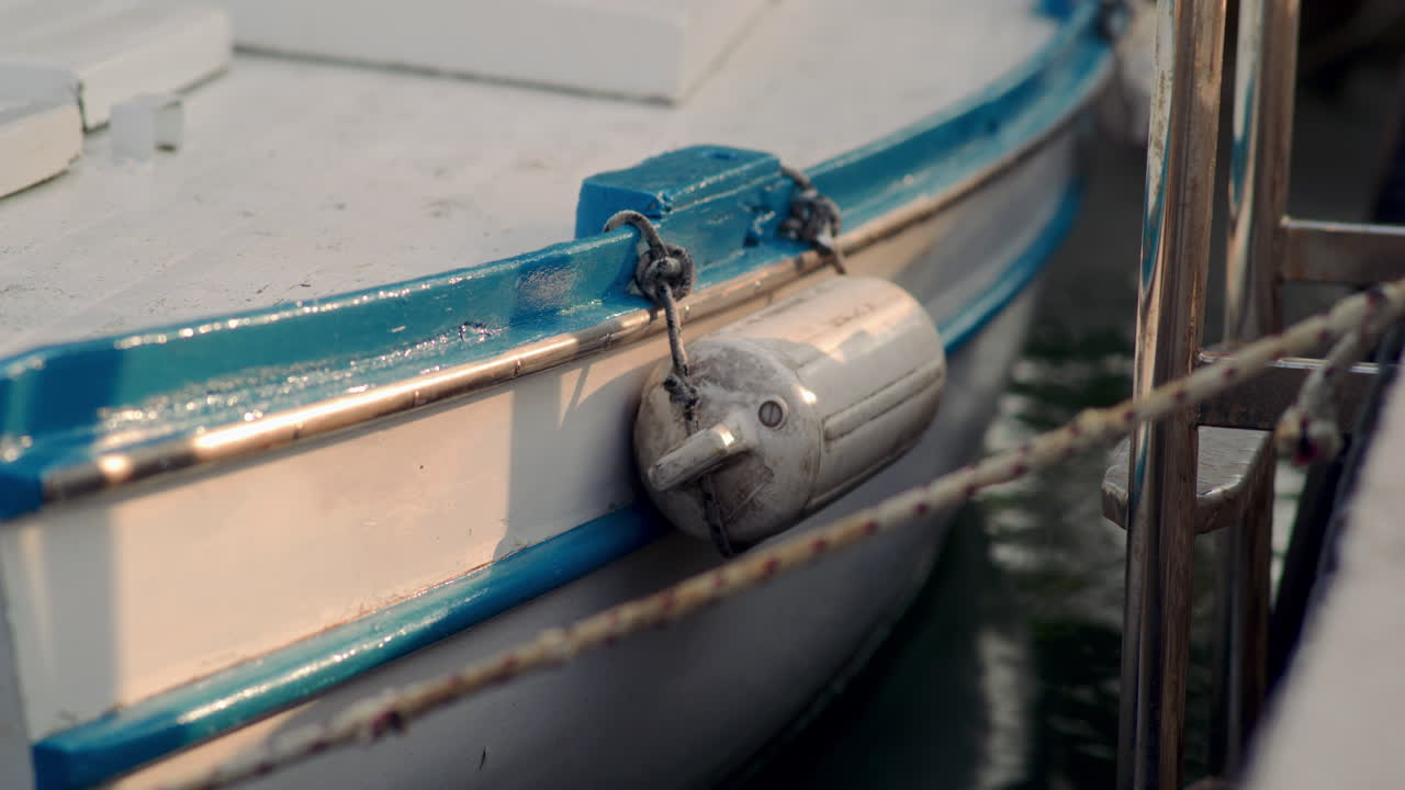 toma detallada de una boya cilíndrica colgada de clips metálicos en el lado de un barco de madera blanco y azul iluminado por la luz de la hora dorada