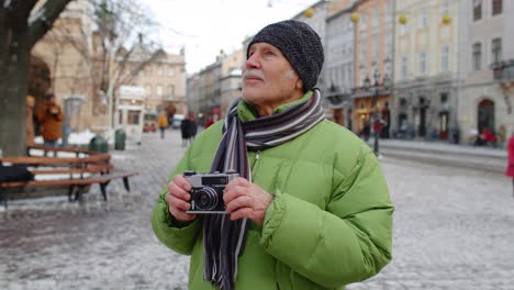 Senior-old-man-taking-pictures-with-photo-camera,-smiling-using-retro-device-in-winter-city-center