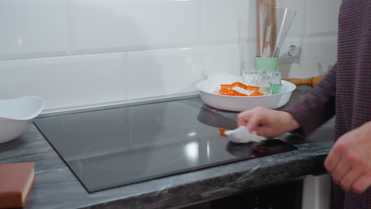 Visuale laterale di una donna che pulisce un elegante piano cottura in vetro in una cucina moderna; la mano tiene un panno e lucida delicatamente la superficie, assicurando la pulizia; piano di lavoro elegante e arredamento minimalista