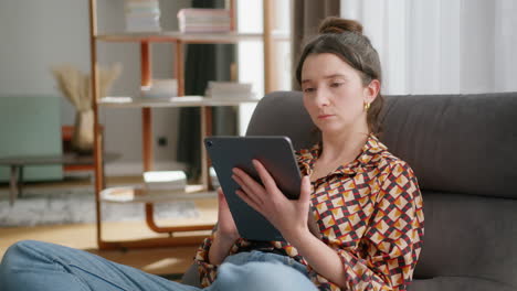 young woman sits on couch in living room and uses tablet, medium shot