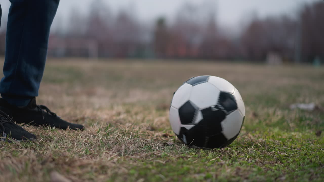 Close-up of a person stopping a rotating soccer ball with his left foot on a grassy field, with a blurred background showing a football goalpost