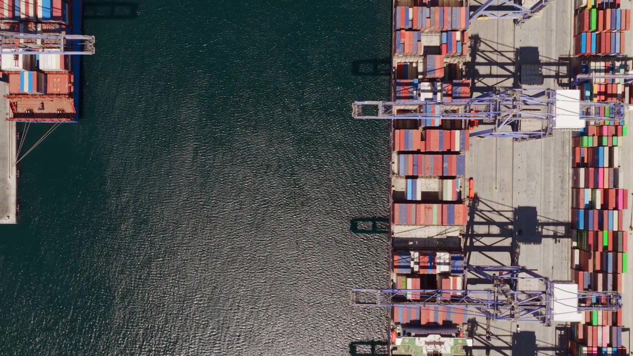 Top-down aerial view showing a large cargo container ship departing from a commercial port terminal, revealing the industrial harbor and rows of colorful intermodal containers on the quay