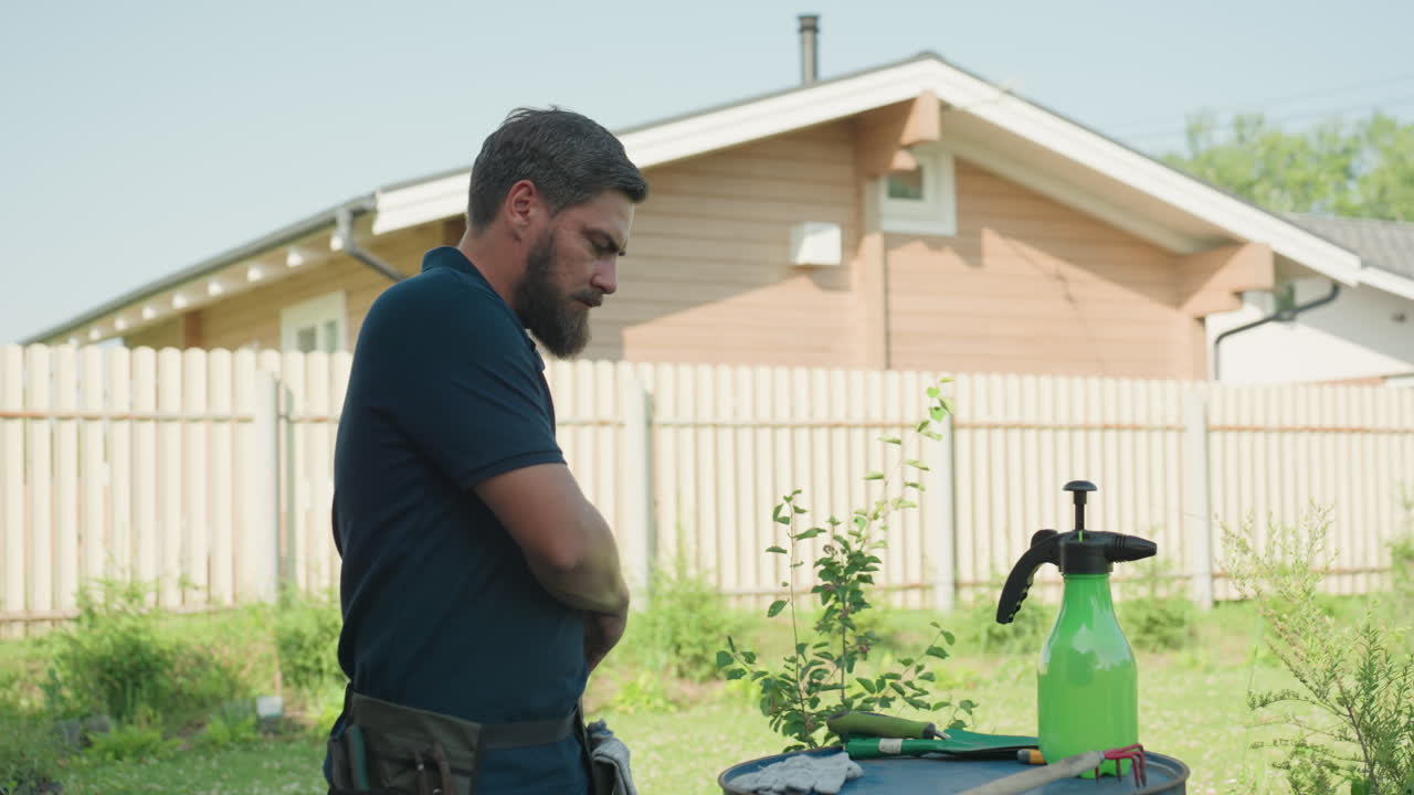 Gardener in yard picking pliers, trowel fork, trowel from drum, placing tools into apron belt around waist beside house sprayer, organizing gear for garden work in sunny summer