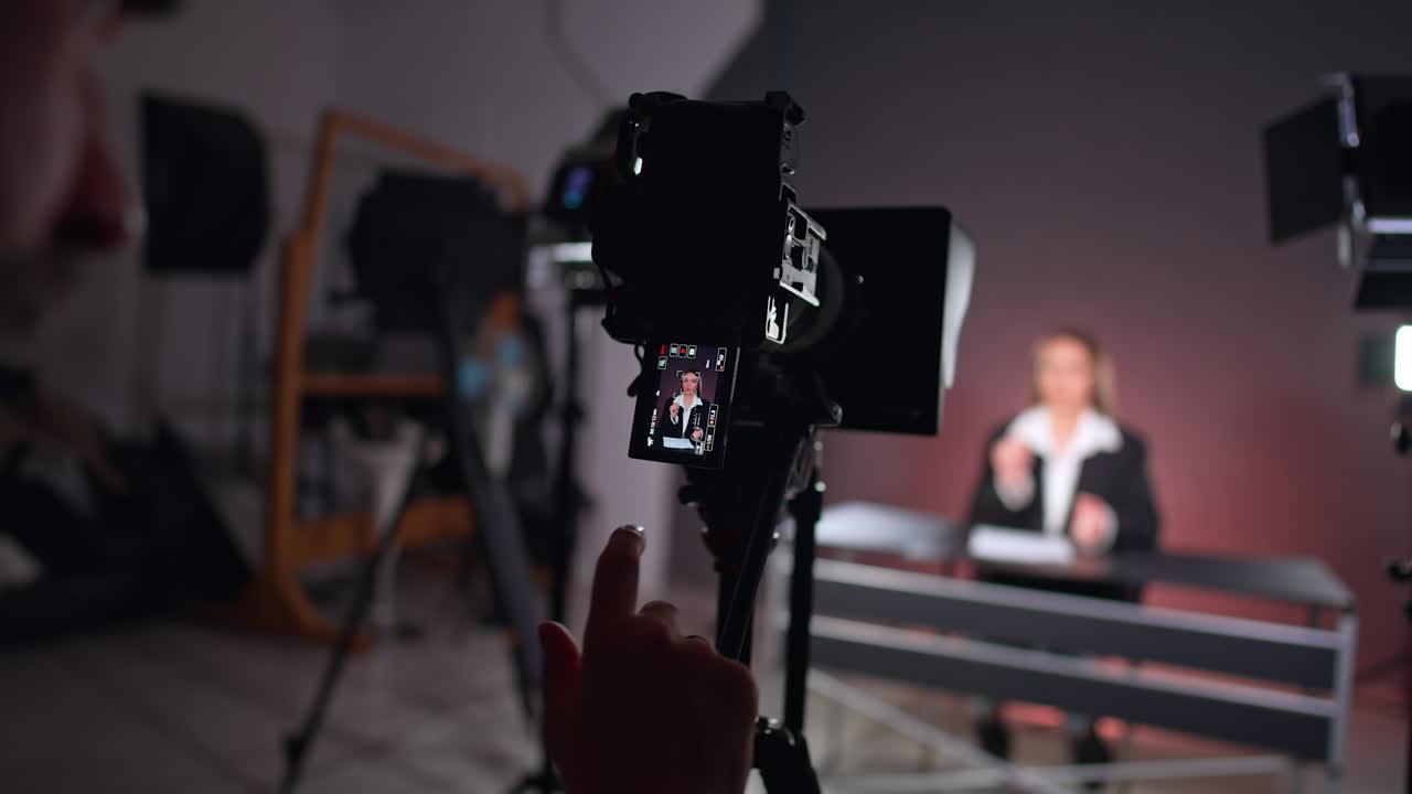 Display of camera on tripod shows the lady in black jacket. Cameraman stands at camera looking at display. Studio footage of blog.