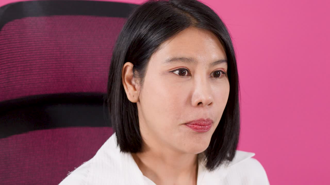 Ảnh cận trung của một người phụ nữ châu Á tươi cười đang nói chuyện và diễn đạt bằng cử chỉ trên nền hồng rực rỡ