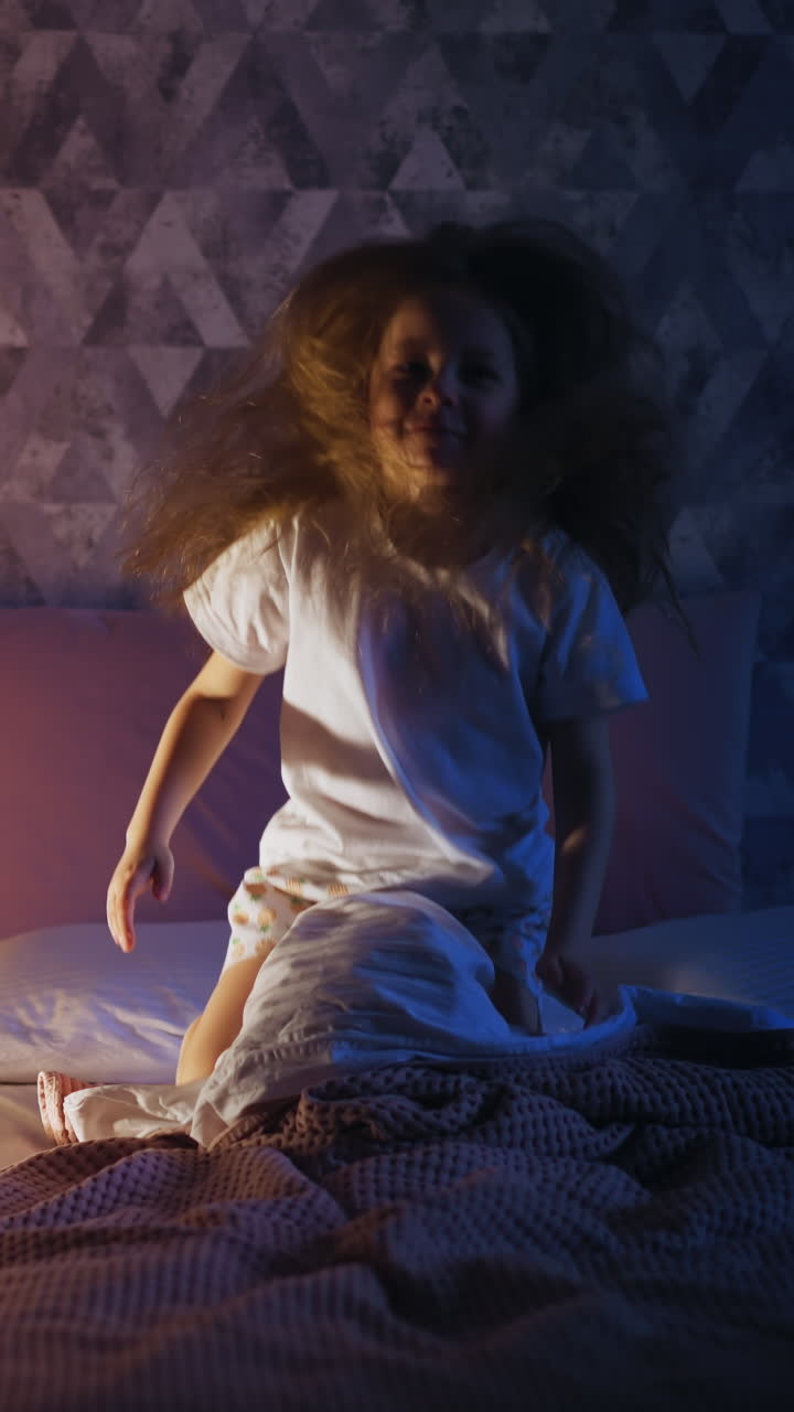 Naughty little girl with loose long hair jumps on large bed near high play tent with glowing night lamp in dark bedroom at home in evening slow motion