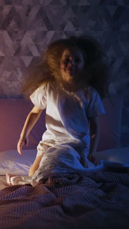 naughty little girl with loose long hair jumps on large bed near high play tent with glowing night lamp in dark bedroom at home in evening slow motion