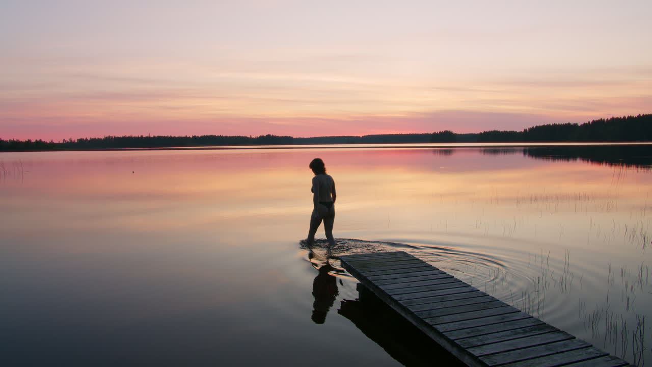 Silhouetted Woman Going Swimming in Beautiful Clear Lake in Tranquil Summer Sunset, Leaving Ripples Behind Her as She Leaves Wooden Pier in Northern Sweden, High Quality Nature Adventure Shot