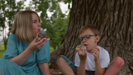 a mother and her son sit under a tree, enjoying a moment together as they eat chocolate cake