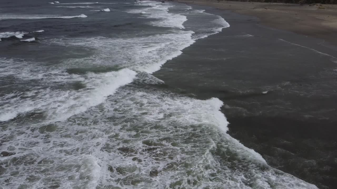 On a gloomy day, the drone camera pans up from the waves battering the beach to reveal lush green hills, rocky cliffs, and trees dotting the landscape. Filmed on a gloomy day in Northern California.