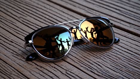 sunglasses with mirrored lenses reflecting silhouettes of people dancing are lying on a weathered wooden surface, creating a contrast between leisure and rustic setting
