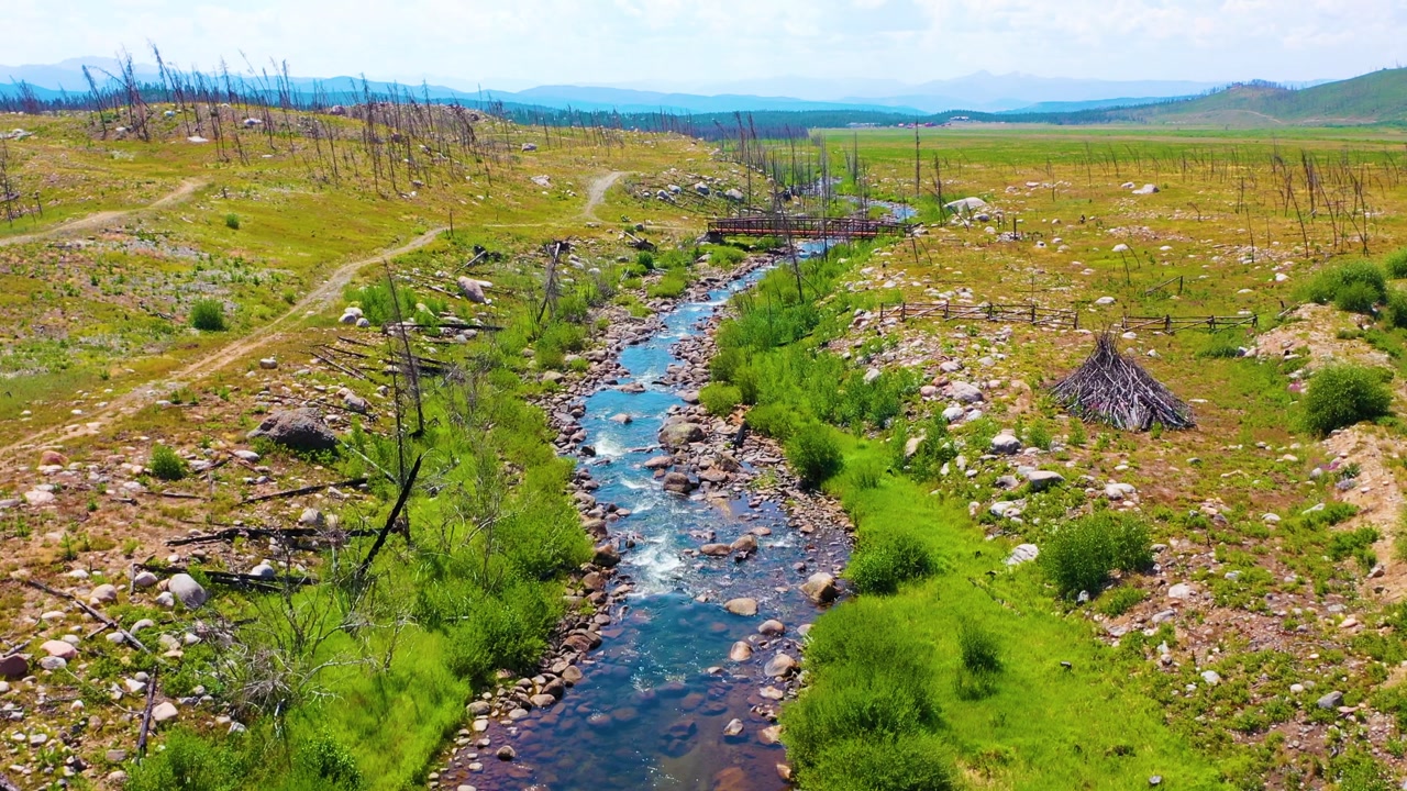 Backward aerial along a Grand Teton style stream in Wyoming with boulder lined water open grassland sparse trees vivid creekside vegetation and layered mountain scenery revealed