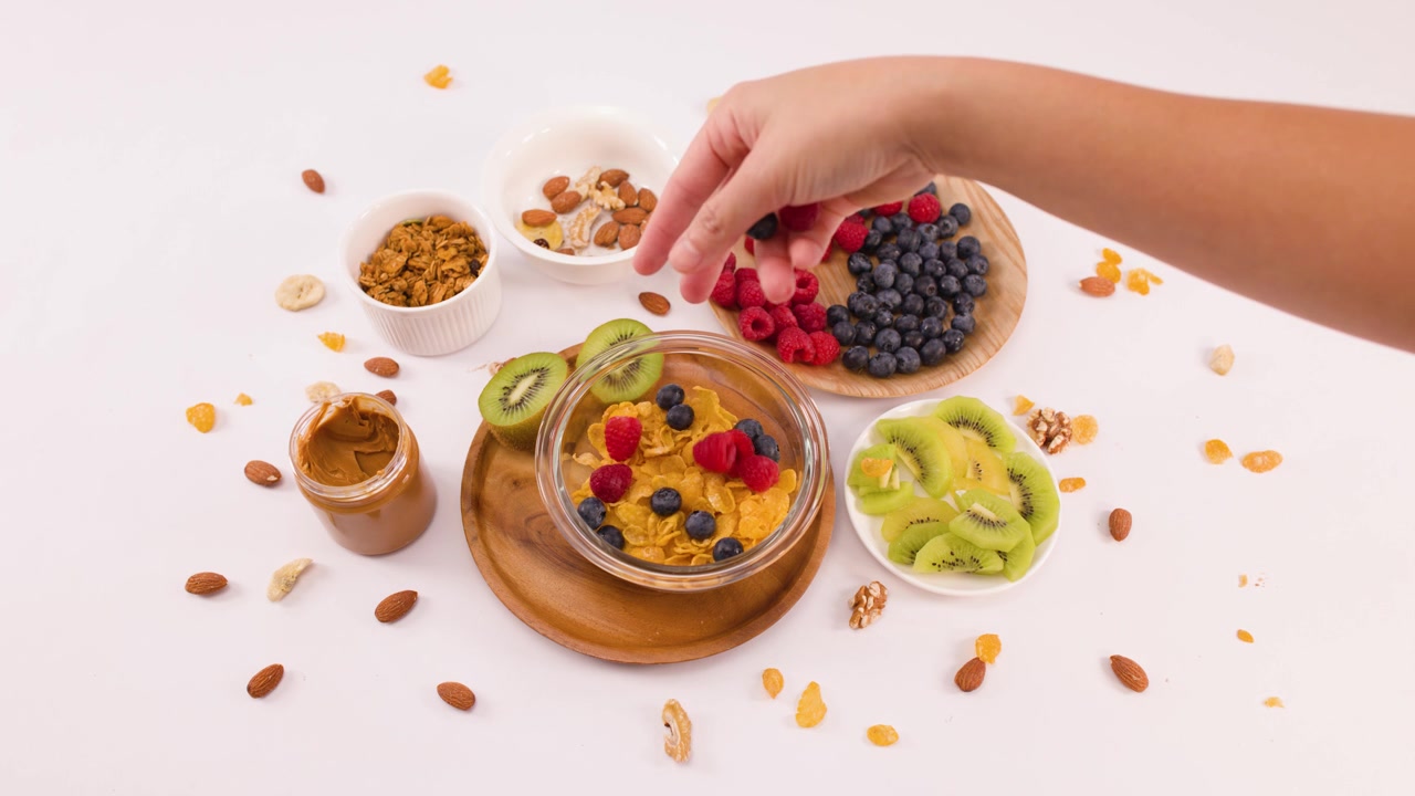 Ripresa dall'alto di una mano che mette lamponi nei cornflakes con frutta, noci e burro di arachidi