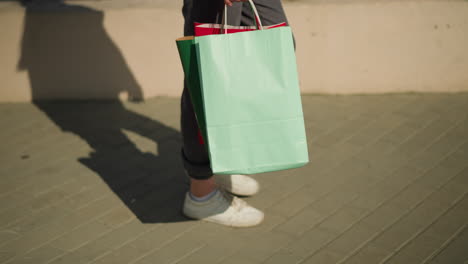 leg view of an individual walking on an interlocked path near a fence while carrying a shopping bag, as the sun casts her shadow while she walks