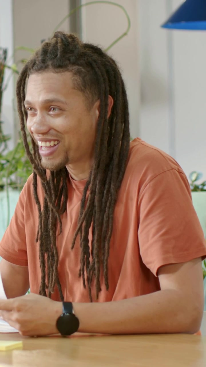 Video verticale: Uomo con i dreadlocks in maglietta arancione che riceve e legge un documento a una riunione, discutendo.