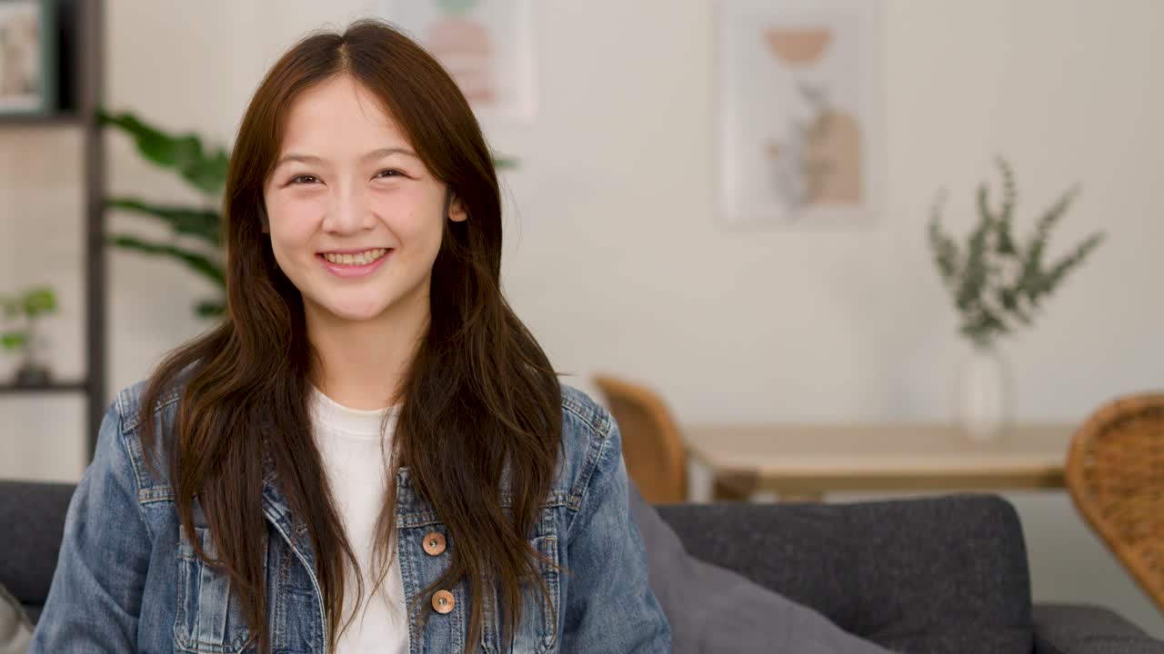 หญิงสาววัยใสในเสื้อแจ็กเก็ตยีนส์ยิ้มอย่างมั่นใจบนโซฟา บรรยากาศสบายๆ ภายในบ้าน