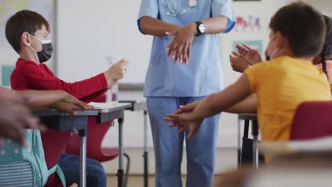 Diverse-schoolchildren-and-medical-worker-wearing-face-masks,-disinfecting-hands-in-classroom