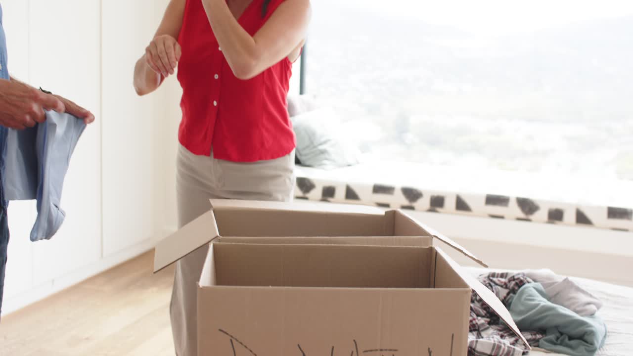 Mature couple sorting and folding light-blue shirts on bed, placing stacks into DONATION box