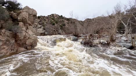 Raging-dirty,-brown-and-muddy-water-rushing-down-Deep-Creek-in-Hesperia,-California-on-an-overcast-day-after-a-massive-flood-with-beautiful-rocky-surroundings