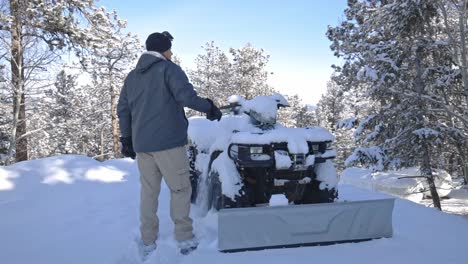 a caucasian male approaching and brushing snow off of an atv or 4 wheeler with a plow to clear snow from a mountain driveway